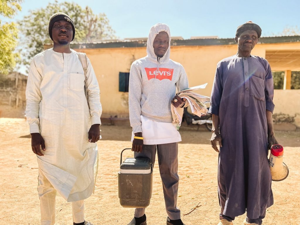 Three men stand facing the camera. The man in the center holds a cooler and sheaf of papers. The man on the right holds a bullhorn.